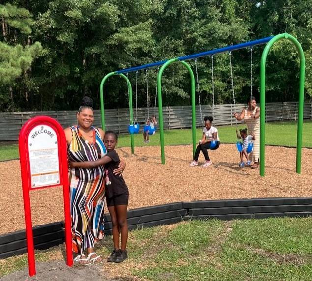 A woman and child standing next to a welcome sign. They are standing in front of a swing set with a woman and three children.