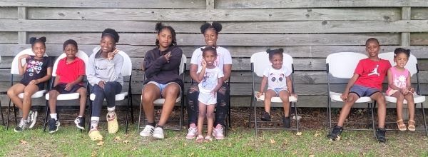 Group of kids in chairs along a wooden wall posing for a photo.