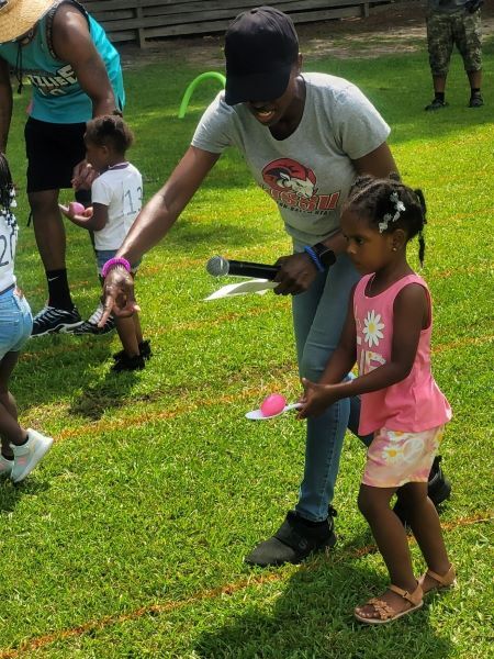 A woman with a microphone helps a young girl with an egg on a spoon in a race. 