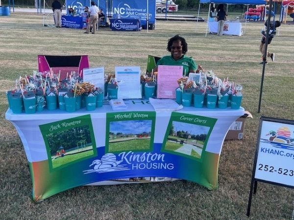 A woman sits at the Kinston Housing table, that has cups filled with goodies.